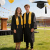 Two young adults in black graduation gowns and caps stand outdoors on a grassy field.
