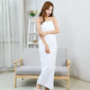 A woman wearing a white, sleeveless maxi dress stands next to a light gray sofa in a bright room.