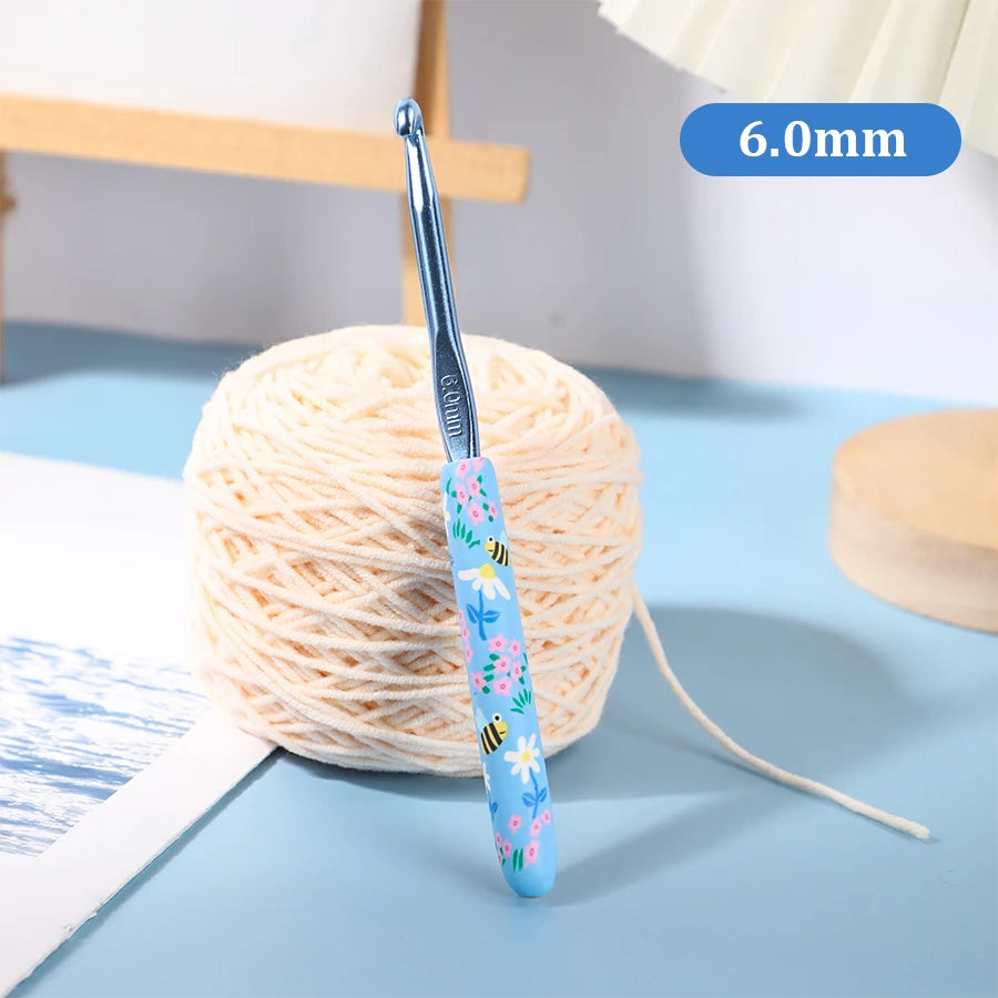 A blue ergonomic crochet hook with a bee design is shown next to a ball of light pink yarn.