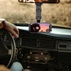 A hand holds a phone mounted on a rotating car rearview mirror phone holder inside a vehicle.