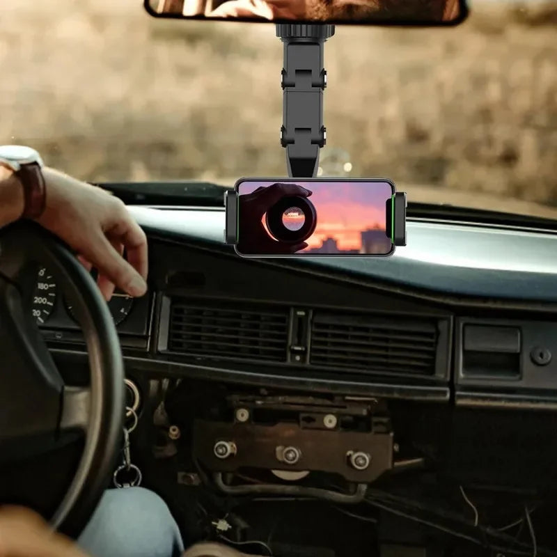 A hand holds a phone mounted on a rotating car rearview mirror phone holder inside a vehicle.