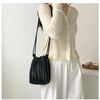 A woman holds a black pleated faux leather crossbody bag against a white background while wearing a cream knit top and white 