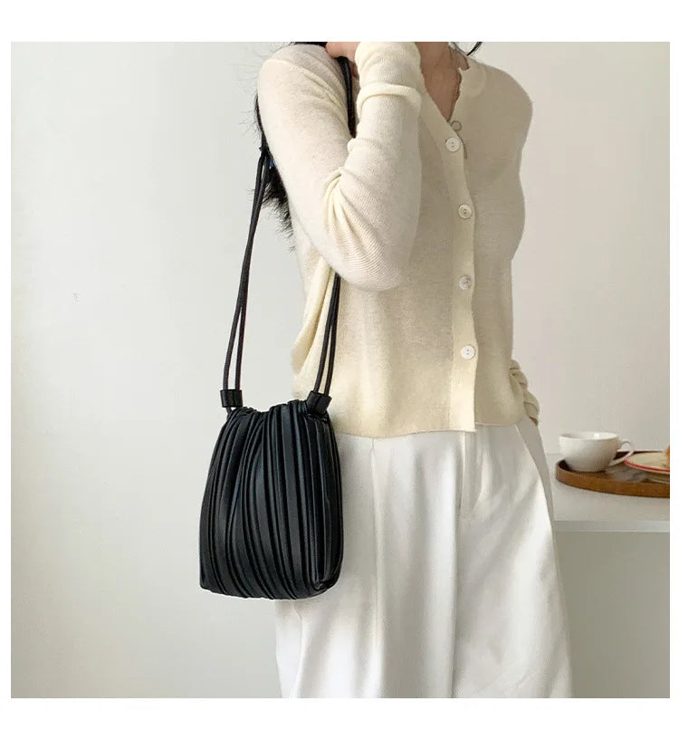 A woman holds a black pleated faux leather crossbody bag against a white background while wearing a cream knit top and white 