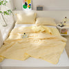 A yellow quilted blanket is spread out on a white bed with pillows and a small potted plant in the background.