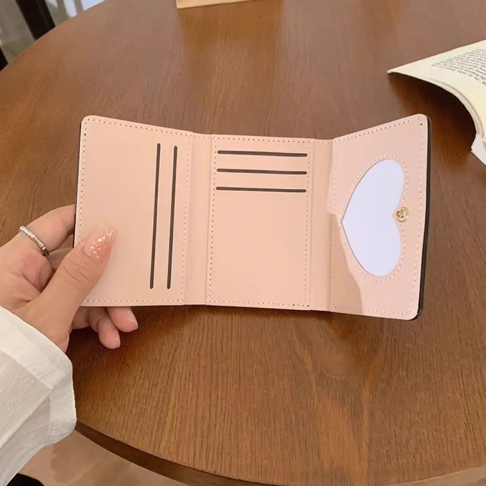 A light pink tri-fold wallet with multiple card slots and a heart cutout is held open on a wooden table.