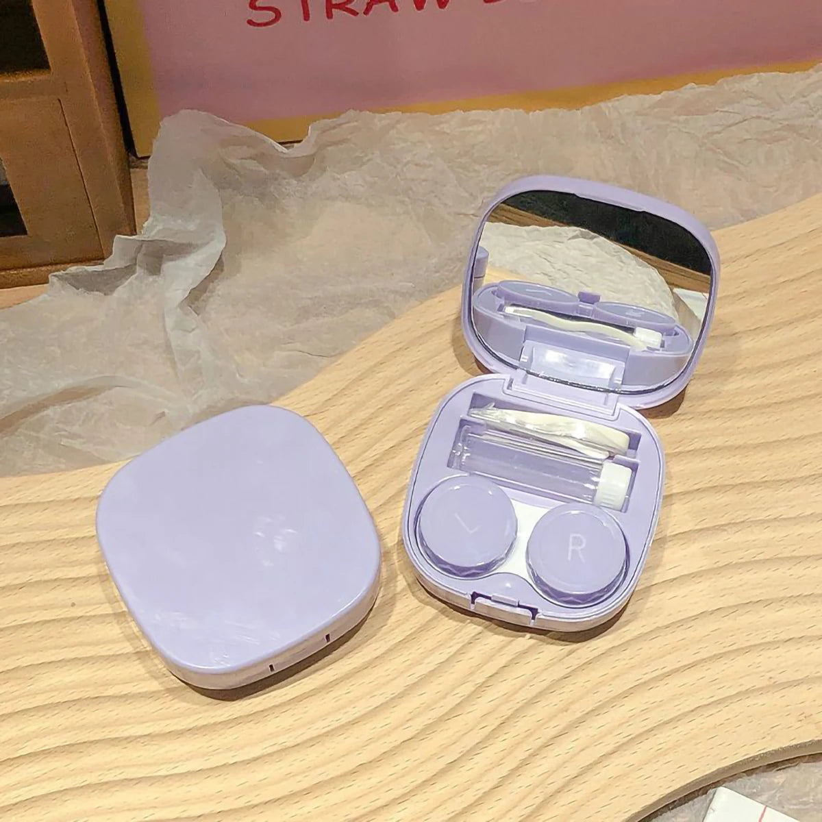 A light purple compact contact lens travel case with a hinged lid is displayed on a wooden surface.