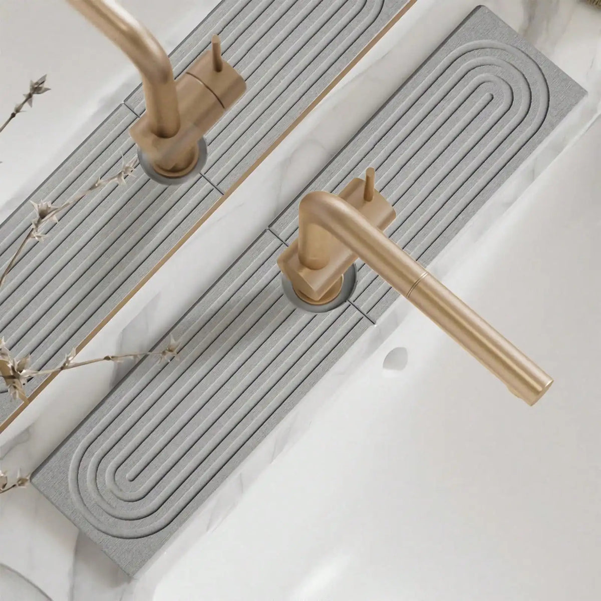 A brass faucet mat with a wavy design is installed on a white countertop next to a sink.