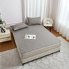 A light gray waterproof mattress protector with elastic straps is displayed on a bed, next to a white upholstered chair and a