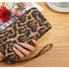 A hand holds a brown leopard print retro long wallet on a wooden table next to some papers and a cup.