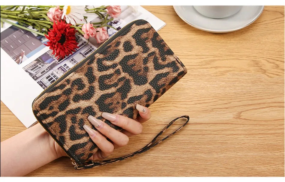 A hand holds a brown leopard print retro long wallet on a wooden table next to some papers and a cup.