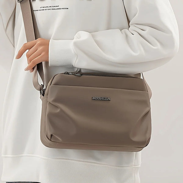 A taupe lightweight nylon crossbody tote bag carried by a person wearing a white sweatshirt.