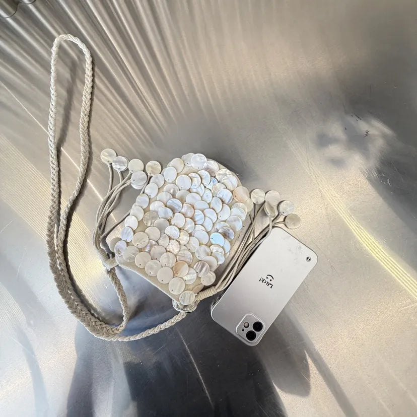 A small shoulder bag adorned with white and light blue sequins and a natural fiber strap, resting on a reflective surface.