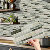 A hand is placing peel-and-stick tile stickers onto a kitchen backsplash made of variegated green and beige stone tiles.