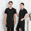 Two models wearing black adjustable cotton aprons standing in front of white paneled doors.