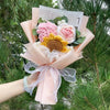 Ein Strauf von handgefertigten Häkelblumen in Rosa- und Gelbtönen, in durchscheinigem rosa Papier eingewickelt.