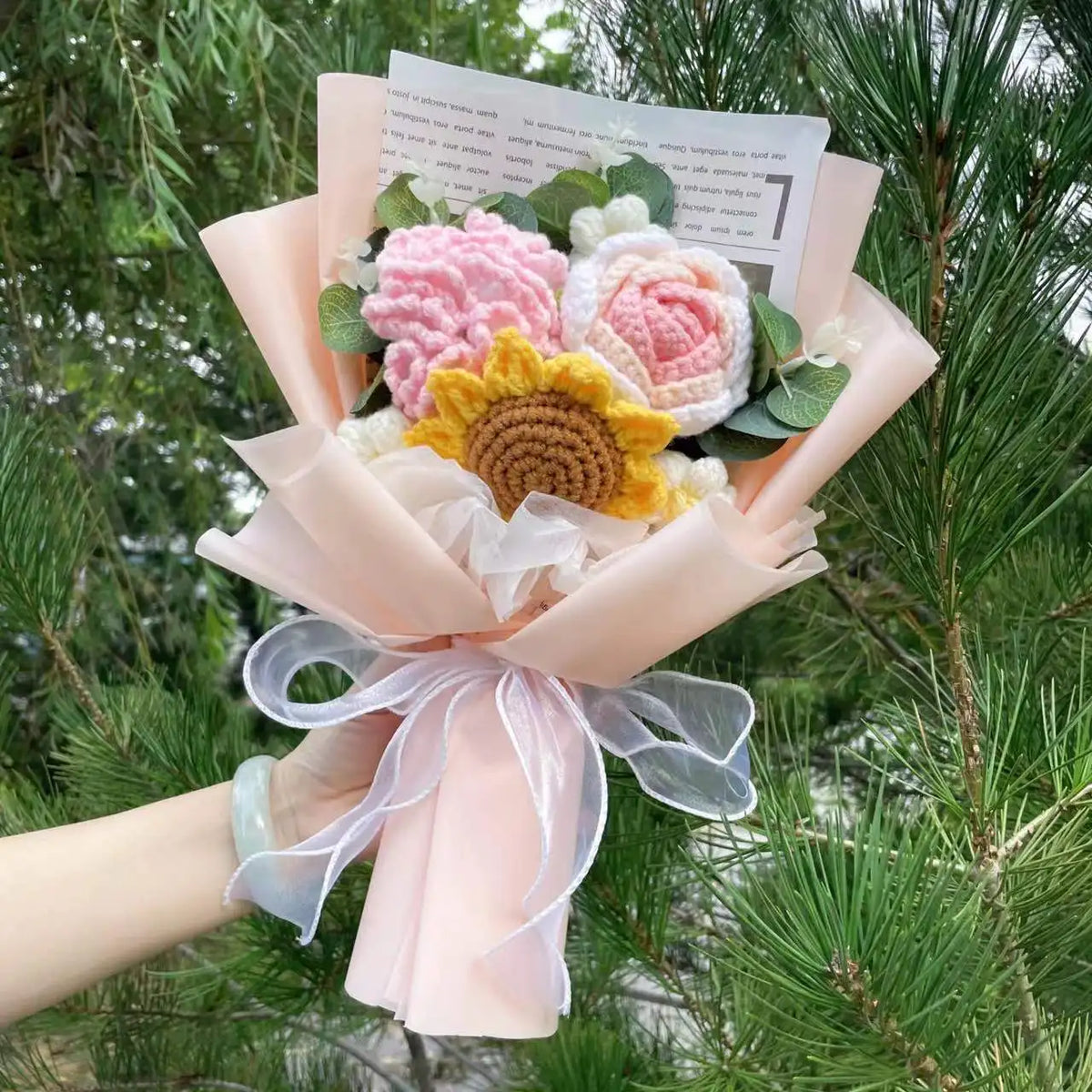 Ein Strauf von handgefertigten Häkelblumen in Rosa- und Gelbtönen, in durchscheinigem rosa Papier eingewickelt.