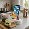 An orange smartphone is mounted on a white base, positioned on a kitchen counter next to a cutting board and vegetables.