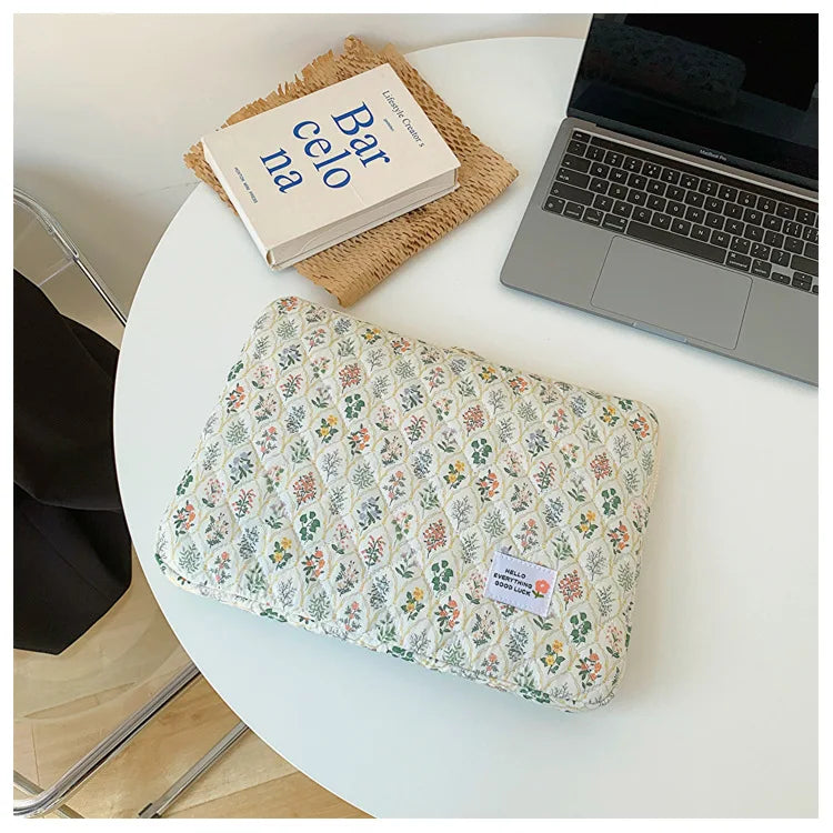 A floral printed laptop sleeve with a zipper closure resting on a white table next to a book and a silver laptop.