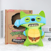 A green and blue plush toy of a turtle monster with large teeth is displayed next to its cardboard packaging.