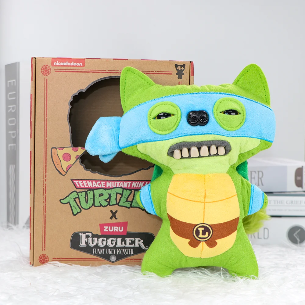 A green and blue plush toy of a turtle monster with large teeth is displayed next to its cardboard packaging.