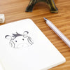 A white A6 kraft paper blank notebook with a simple black line drawing of a horned animal on the cover, next to a silver pen.