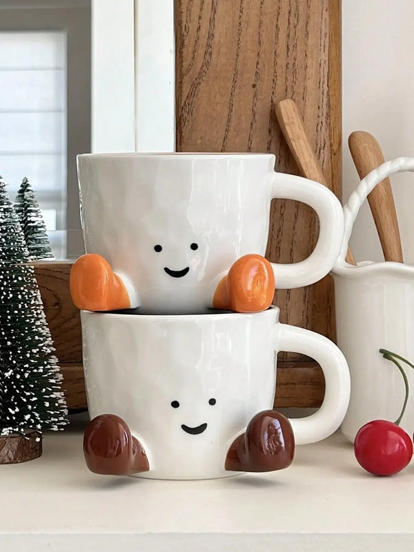 Tasse à café en céramique blanche avec visage souriant et accents orange, posée sur une surface claire.