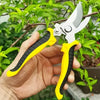 A hand holds a pair of yellow and black stainless steel bypass pruning shears against a green plant background.