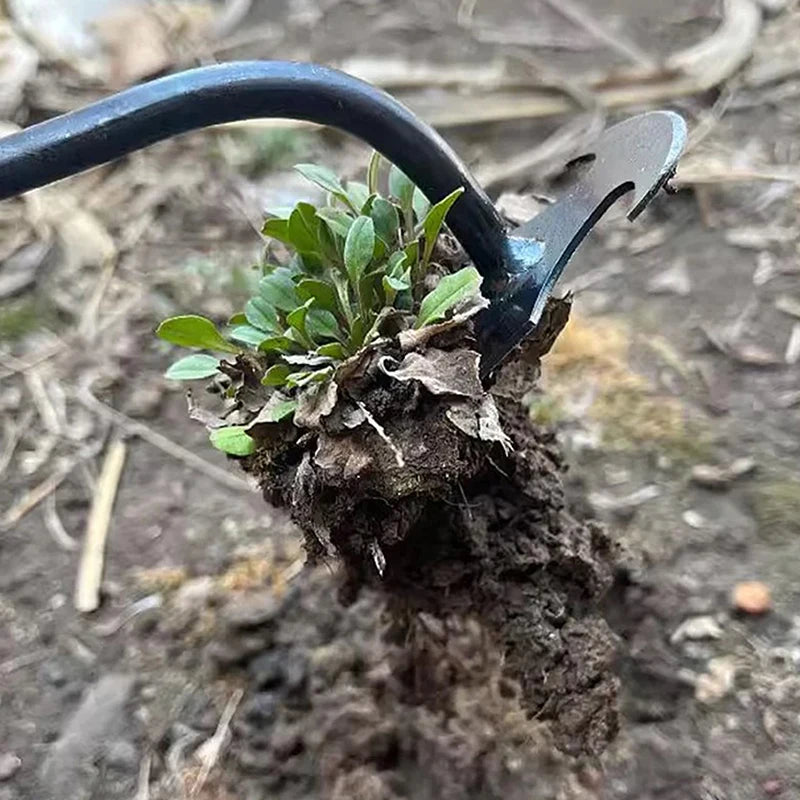 A close-up of a black hand weeder tool with a curved handle, positioned in dark soil near a small green plant.