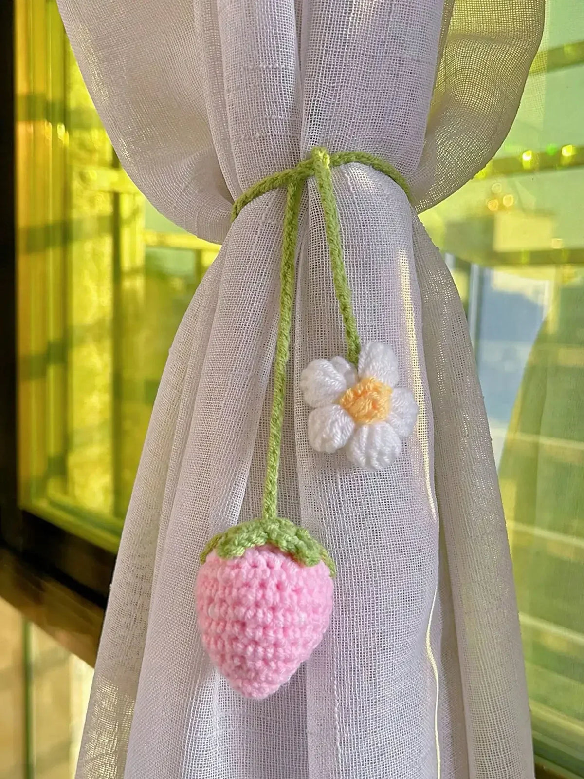 Handmade crochet strawberry pendant with a white flower detail hanging from sheer lilac fabric.