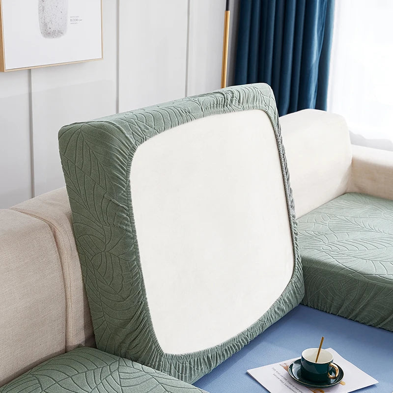 A close-up of a sage green stretch jacquard sofa cover with an anti-slip backing and white cushion on a light-colored sofa.