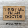 A rectangular tan woven patch with black text that reads TRUST ME I'M AN DOCTOR on a light wooden surface.