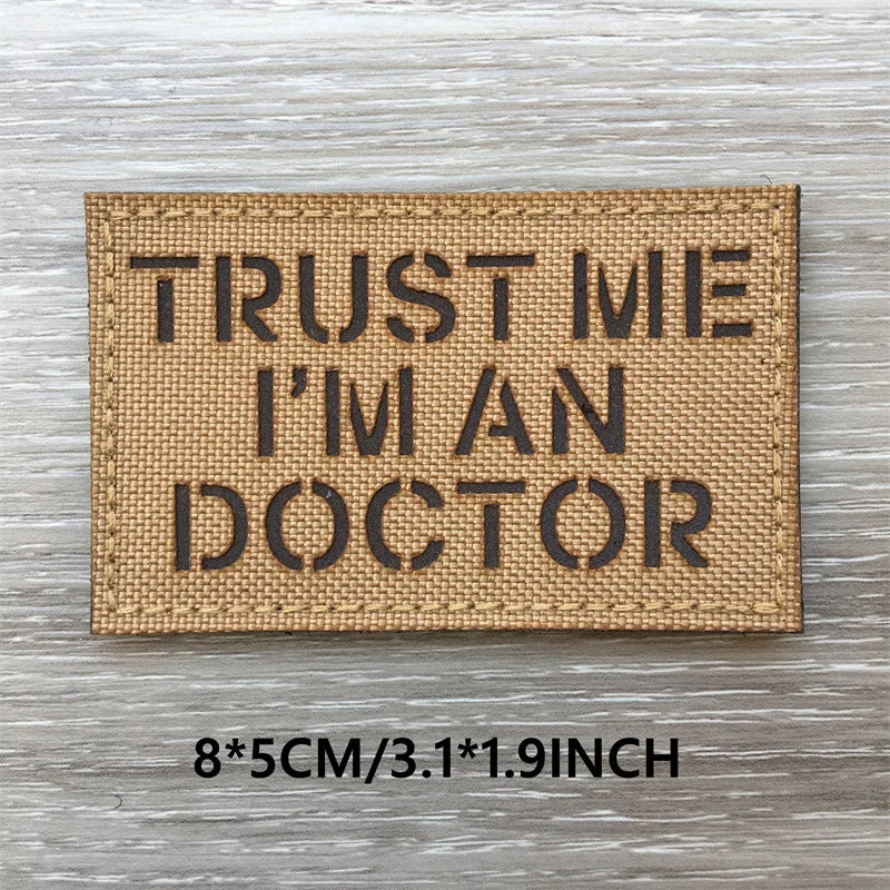 A rectangular tan woven patch with black text that reads TRUST ME I'M AN DOCTOR on a light wooden surface.