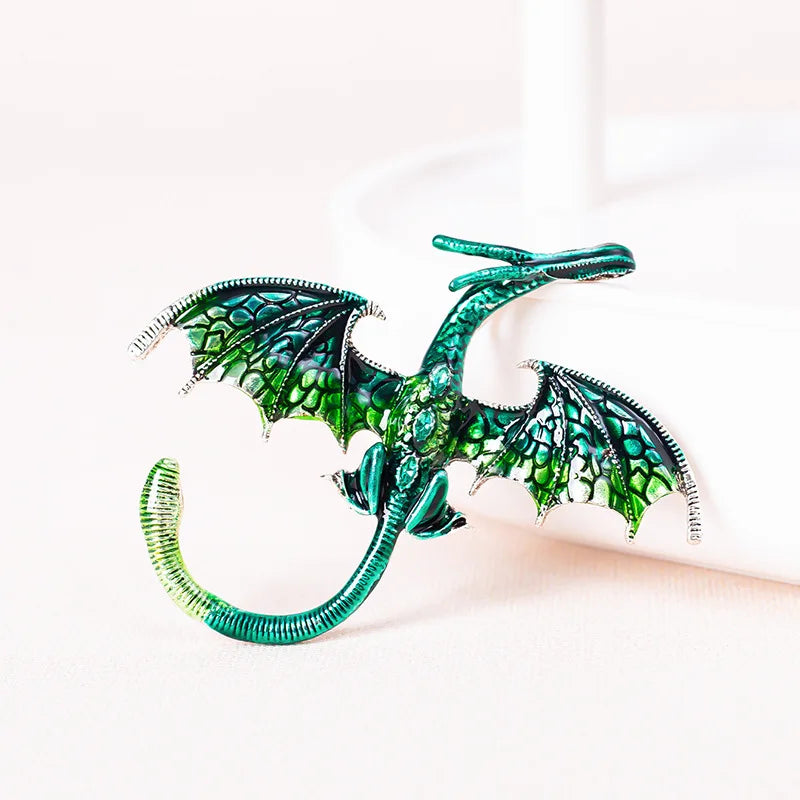 A green enamel dragon brooch with rhinestone accents is displayed on a white surface next to a white candle.