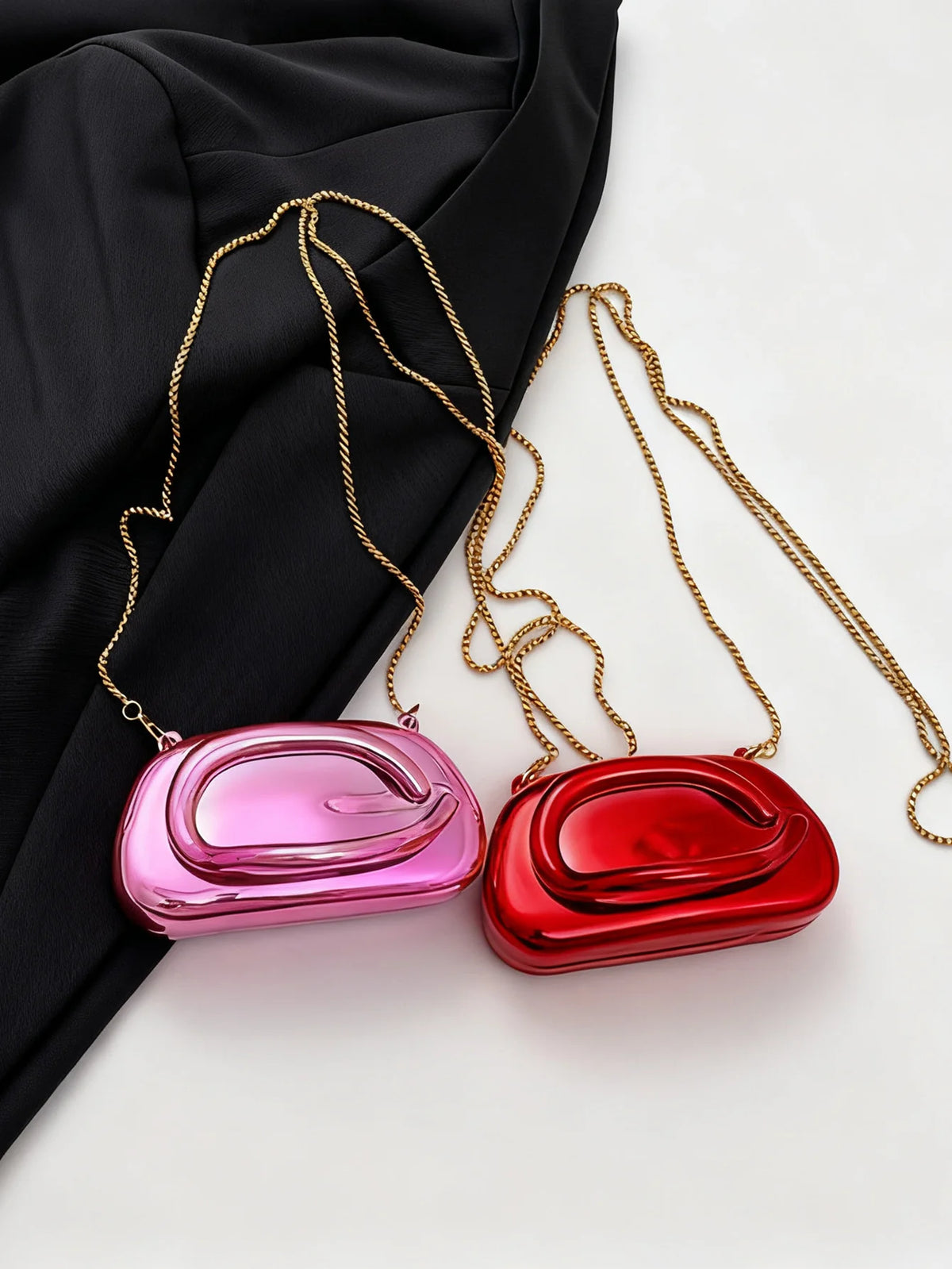A pair of mini gold metal clutch bags with long chains are displayed against a black fabric background.