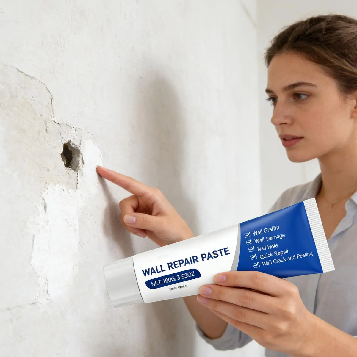 A woman points to a small hole in a white wall while holding a tube of Wall Repair Paste.