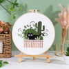 A circular embroidery hoop displaying a pre-printed design of a cactus and a black cat in a floral planter.