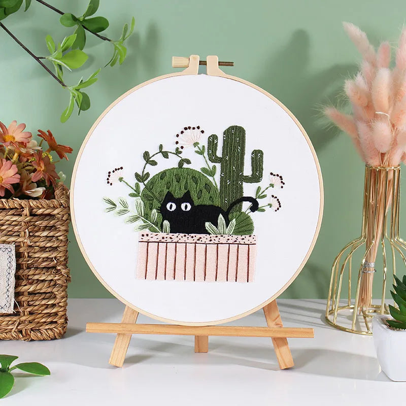 A circular embroidery hoop displaying a pre-printed design of a cactus and a black cat in a floral planter.