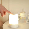 A hand points toward a white LED table lamp with a warm glow next to a glass pitcher.