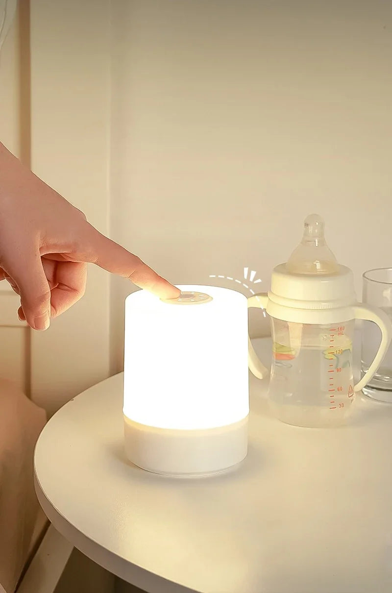 A hand points toward a white LED table lamp with a warm glow next to a glass pitcher.