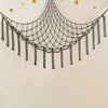 A bohemian plush toy storage hammock made of dark cord hangs against a light wall with gold stars.