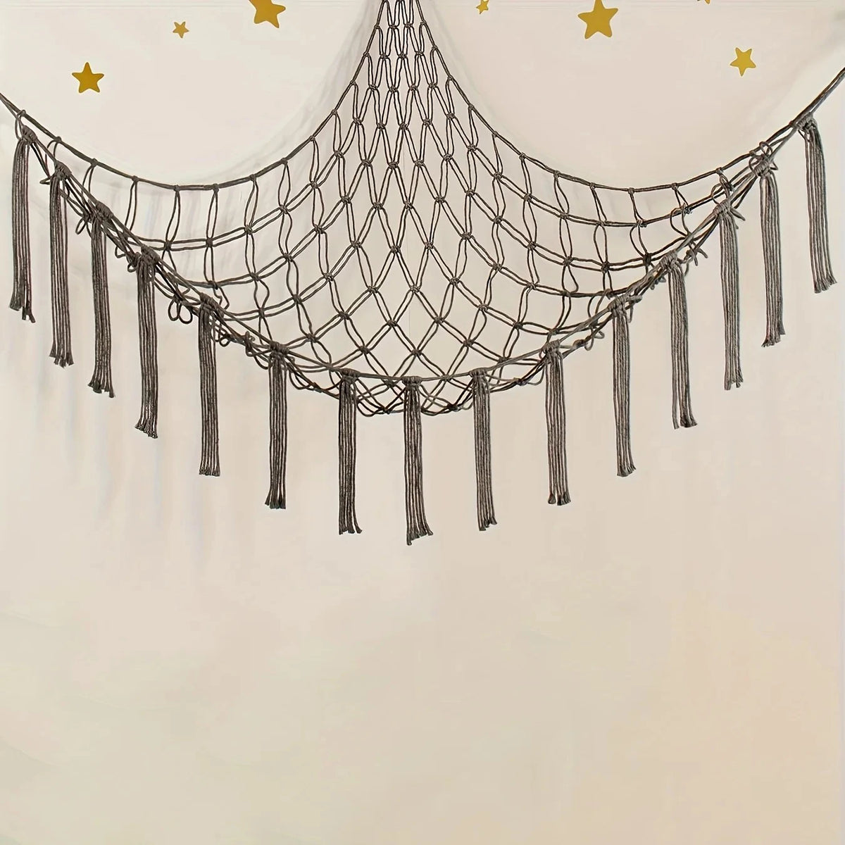 A bohemian plush toy storage hammock made of dark cord hangs against a light wall with gold stars.