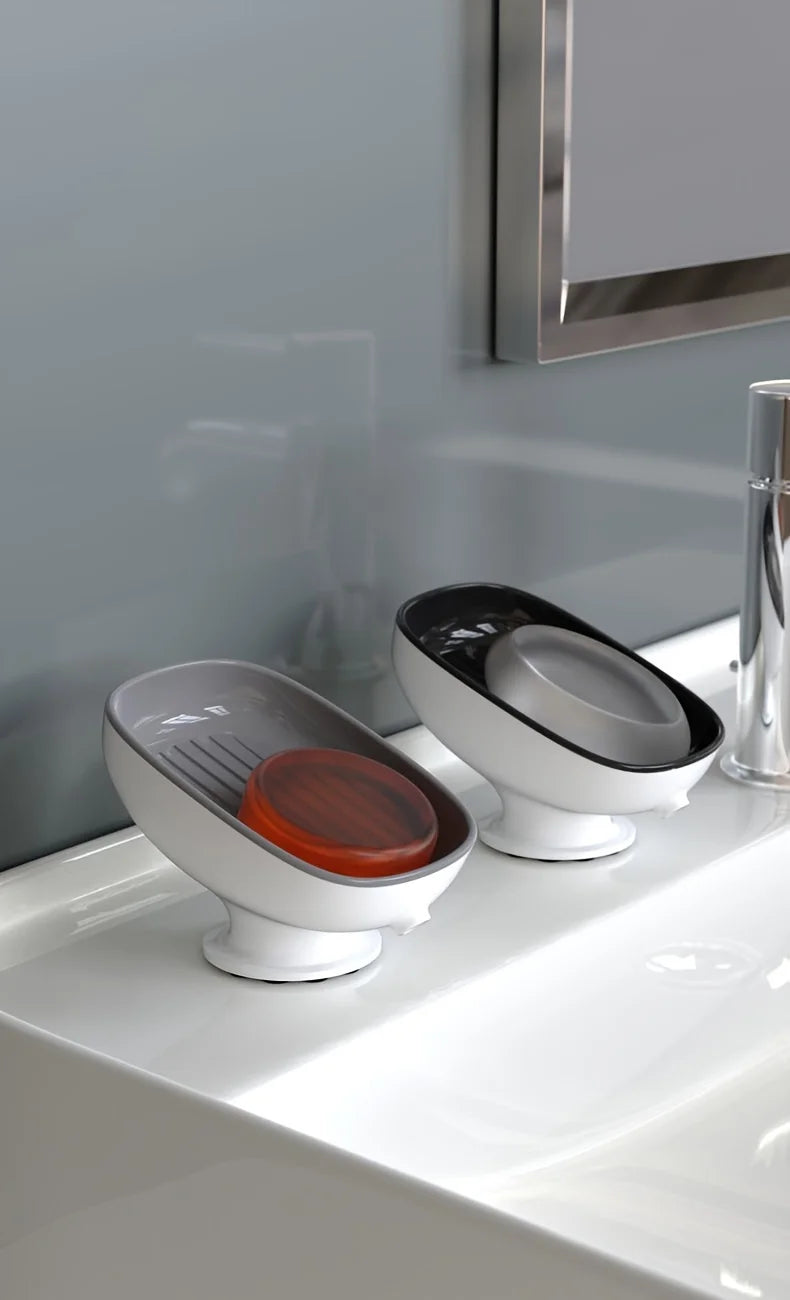 A white soap dish with a leaf-shaped suction cup holder and an orange soap dish sitting on a white bathroom sink.