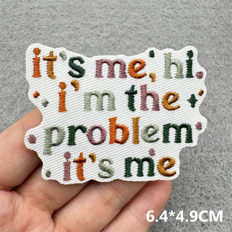 A hand holds a white embroidered patch with text reading it's me hi i'm the problem it's me