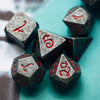 A close-up of seven dark gray metal polyhedral dice with a red dragon font scattered on a teal surface.