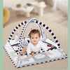 A baby is playing in a white and black striped play gym mat with hanging toys on a light wood floor.