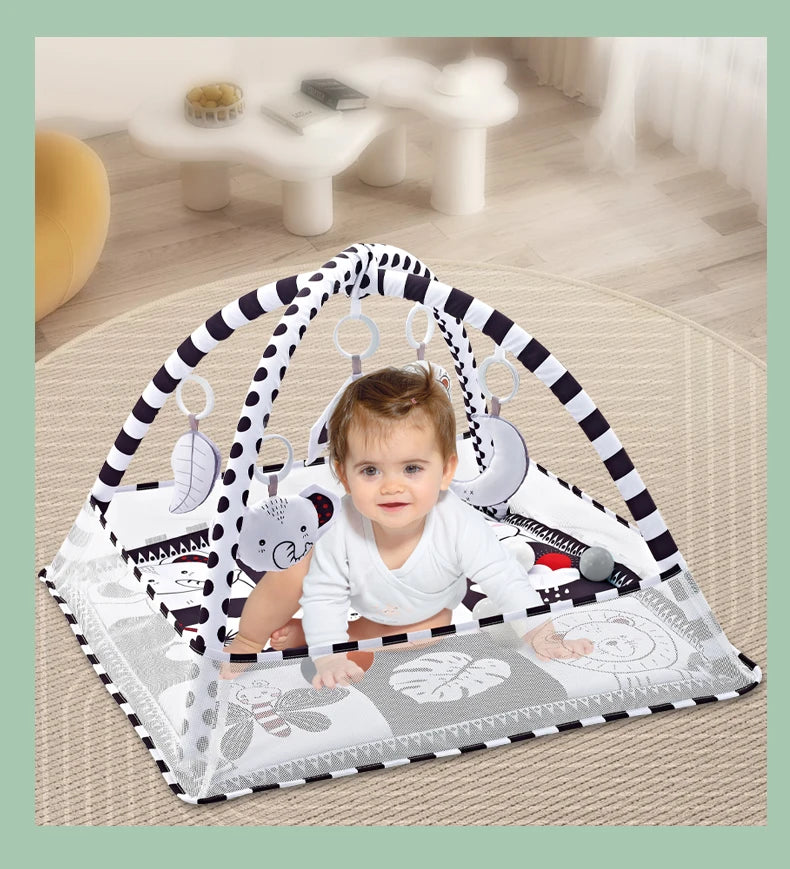 A baby is playing in a white and black striped play gym mat with hanging toys on a light wood floor.