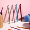 A collection of colorful Tunisian crochet hooks, including green, purple, and red, displayed against a pink background.