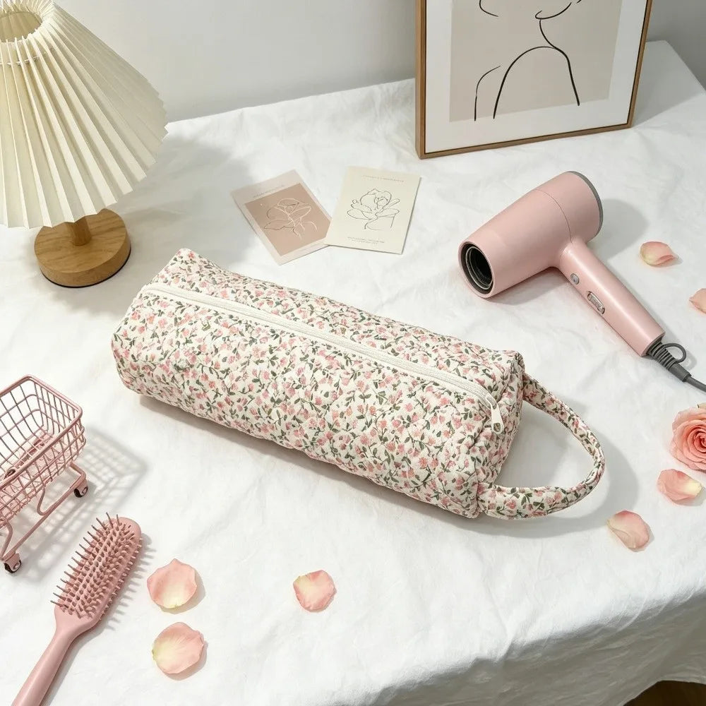 A pink quilted cotton hair dryer storage bag with a handle rests on a white cloth next to a pink hairdryer and a comb.