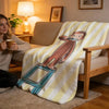 A yellow and white striped throw blanket featuring a cartoon illustration of a man sitting on a blue stool.