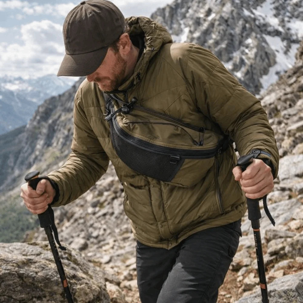 A man wearing a olive green jacket is hiking on rocky terrain while carrying an everyday nylon crossbody bag.